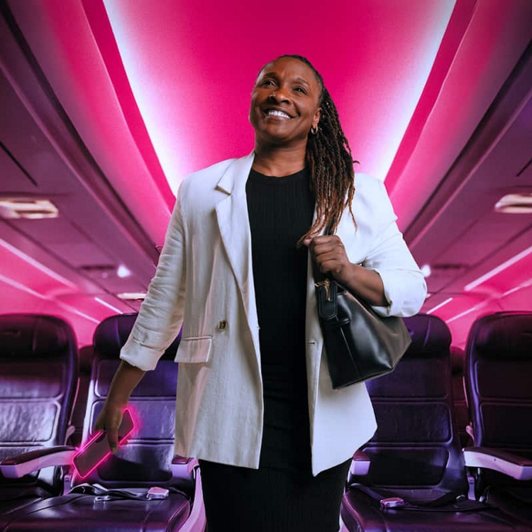 Un viajante de empresas sonriente camina por el pasillo del avión iluminado en magenta con el smartphone en la mano.