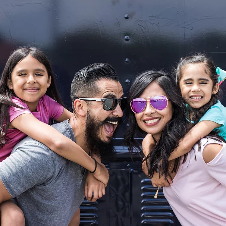 Familia de cuatro sonriendo, con una hija en la espalda del papá y una hija en la espalda de la mamá.